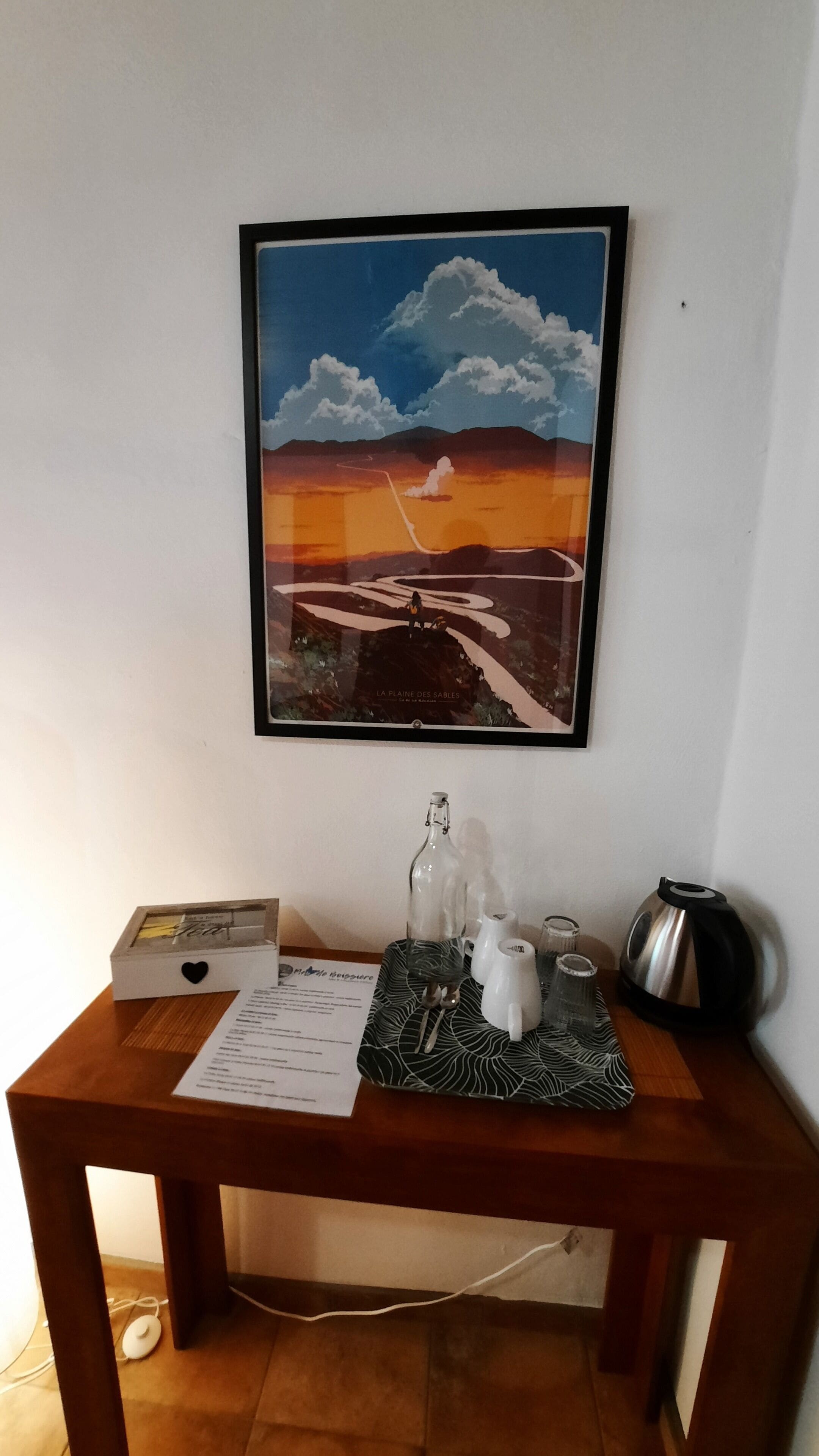 Un cuadro enmarco de un paisaje con una puesta de sol sobre un valle, una pequeña caja blanca con un corazón, una bandeja con tetera, tazas y hervidor, y un menú sobre una mesa de madera.