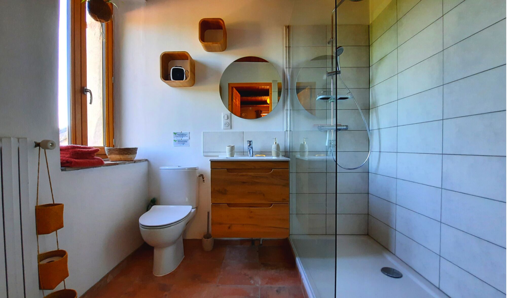 A bathroom with a toilet, sink, and shower. There are wooden shelves above the toilet and a mirror above the sink.