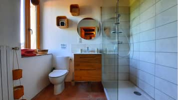 Un bagno con un water, un lavandino e una doccia. Ci sono scaffali in legno sopra il water e uno specchio sopra il lavandino.