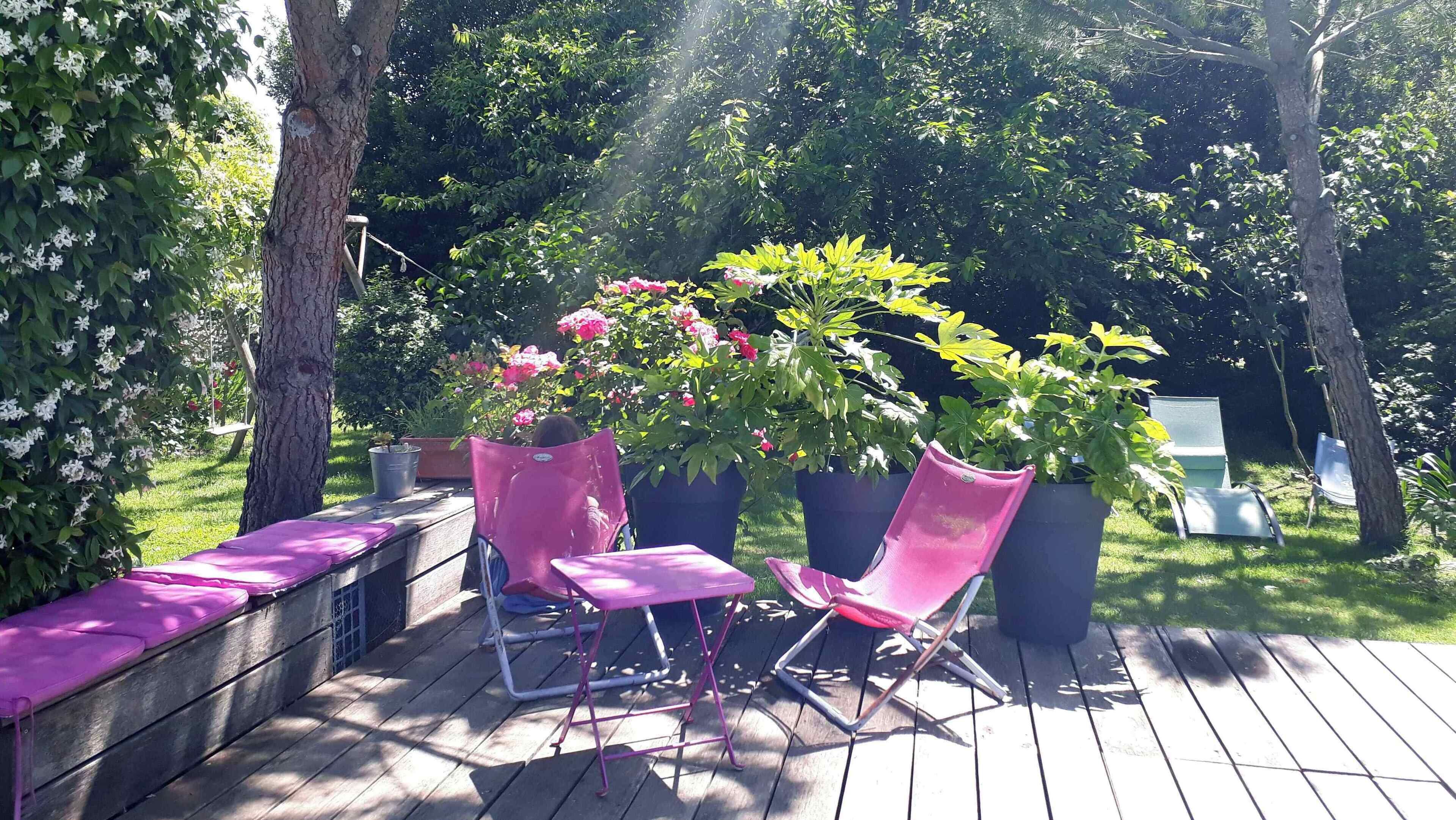 Une terrasse en bois avec deux chaises roses et un banc, entourée d’une végétation luxuriante et de plantes en fleurs.