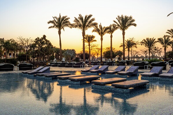 Área de piscina com espreguiçadeiras e palmeiras ao pôr do sol.
