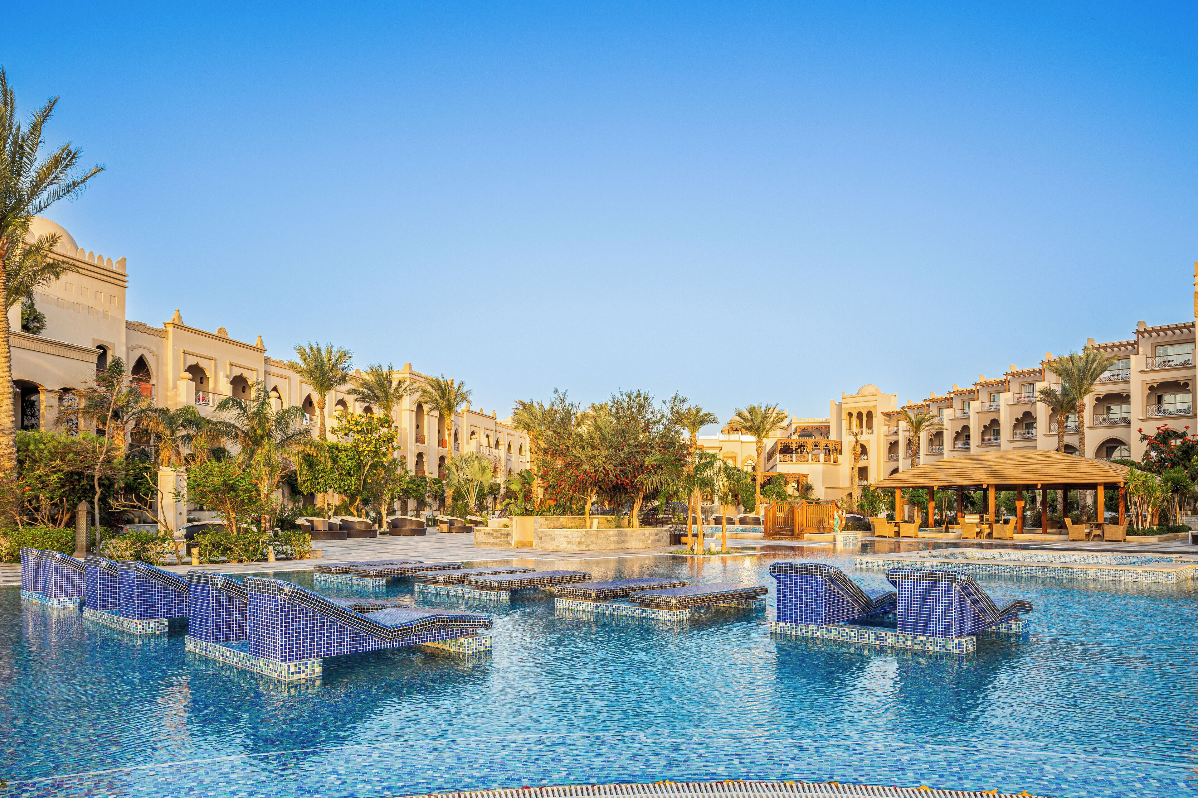 A large pool with blue-tiled steps and lounge chairs, surrounded by palm trees and buildings with arched windows and balconies.
