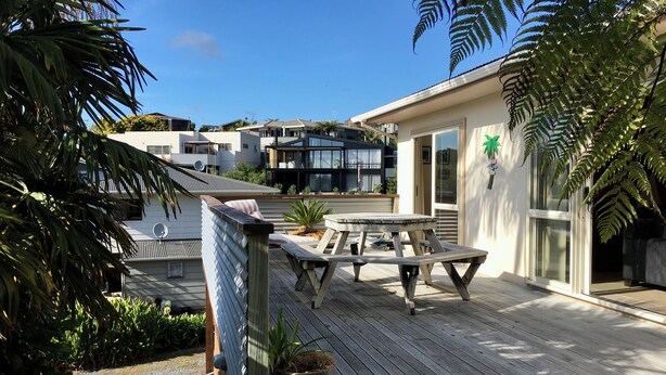 A wooden deck with a picnic table and potted plants, surrounded by modern buildings and a clear blue sky. - Classic 2-bedroom bach in Waihi Beach perfect for relaxing getaway (Waihi Beach)