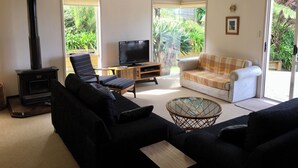 A living room with a black sofa, a white armchair, a television, and a wood-burning stove. - Classic 2-bedroom bach in Waihi Beach perfect for relaxing getaway (Waihi Beach)