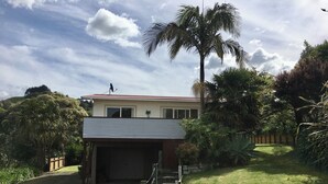 A two-story house with a garage, surrounded by lush greenery and a palm tree. - Classic 2-bedroom bach in Waihi Beach perfect for relaxing getaway (Waihi Beach)