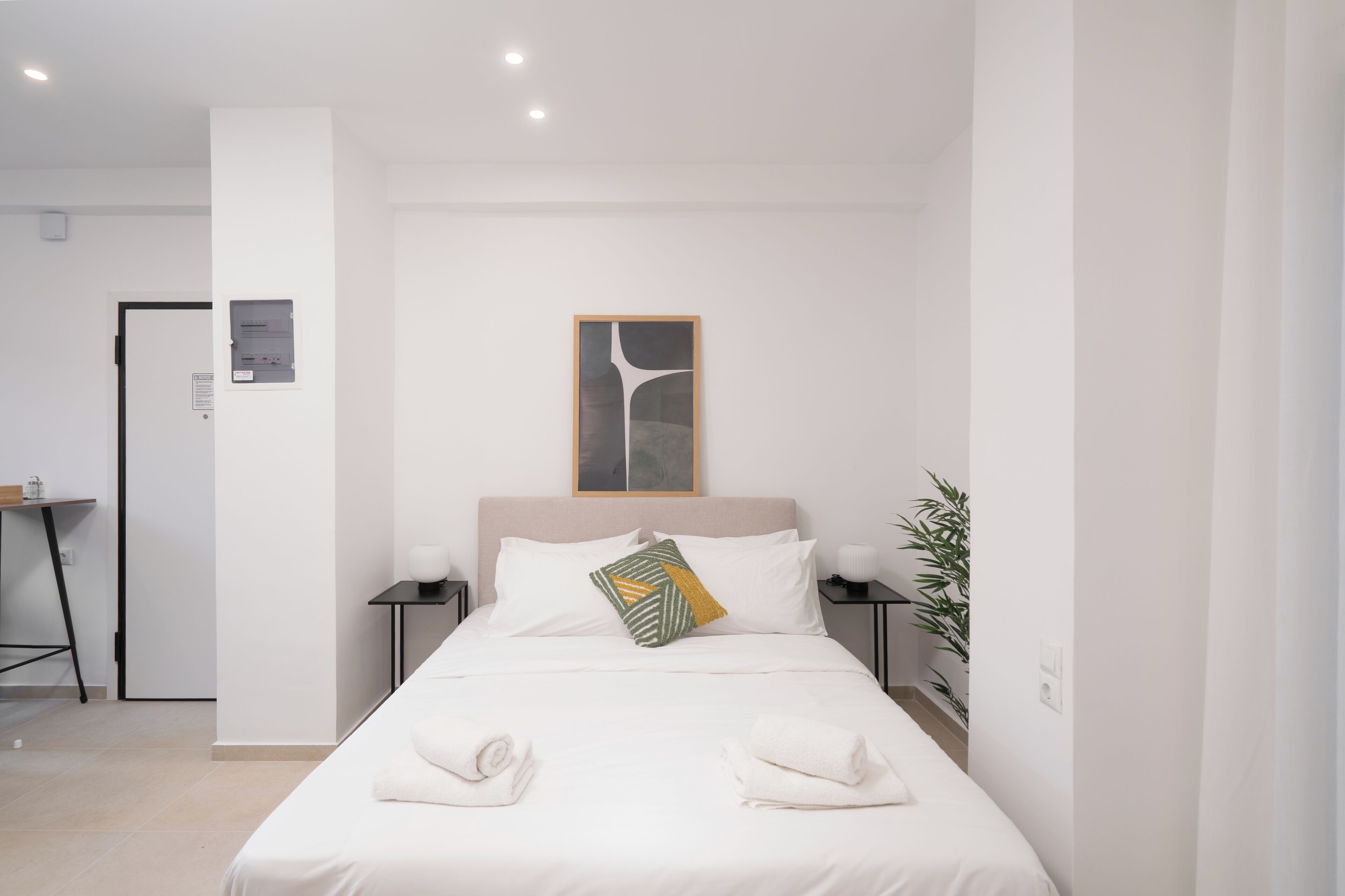 A modern hotel room with a neatly made bed, a bedside table with a lamp, a wall-mounted power outlet, and a door leading to another room.
