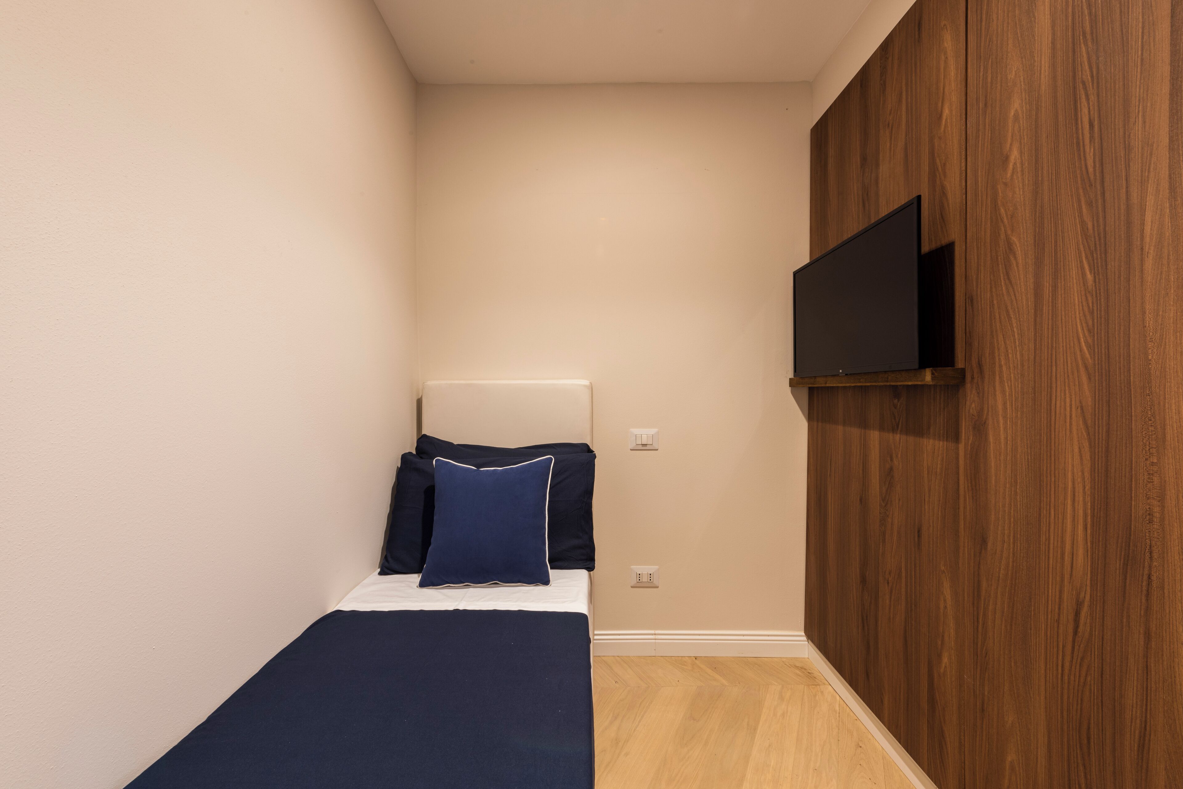 Une petite chambre à coucher, bien rangée, avec un lit, un téléviseur à écran plat fixé au mur et un panneau en bois sur l’un des murs.