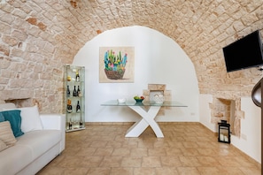 A living room with a stone wall, a glass table, a white sofa, a television, and a fireplace. - Helios Room (Conversano)