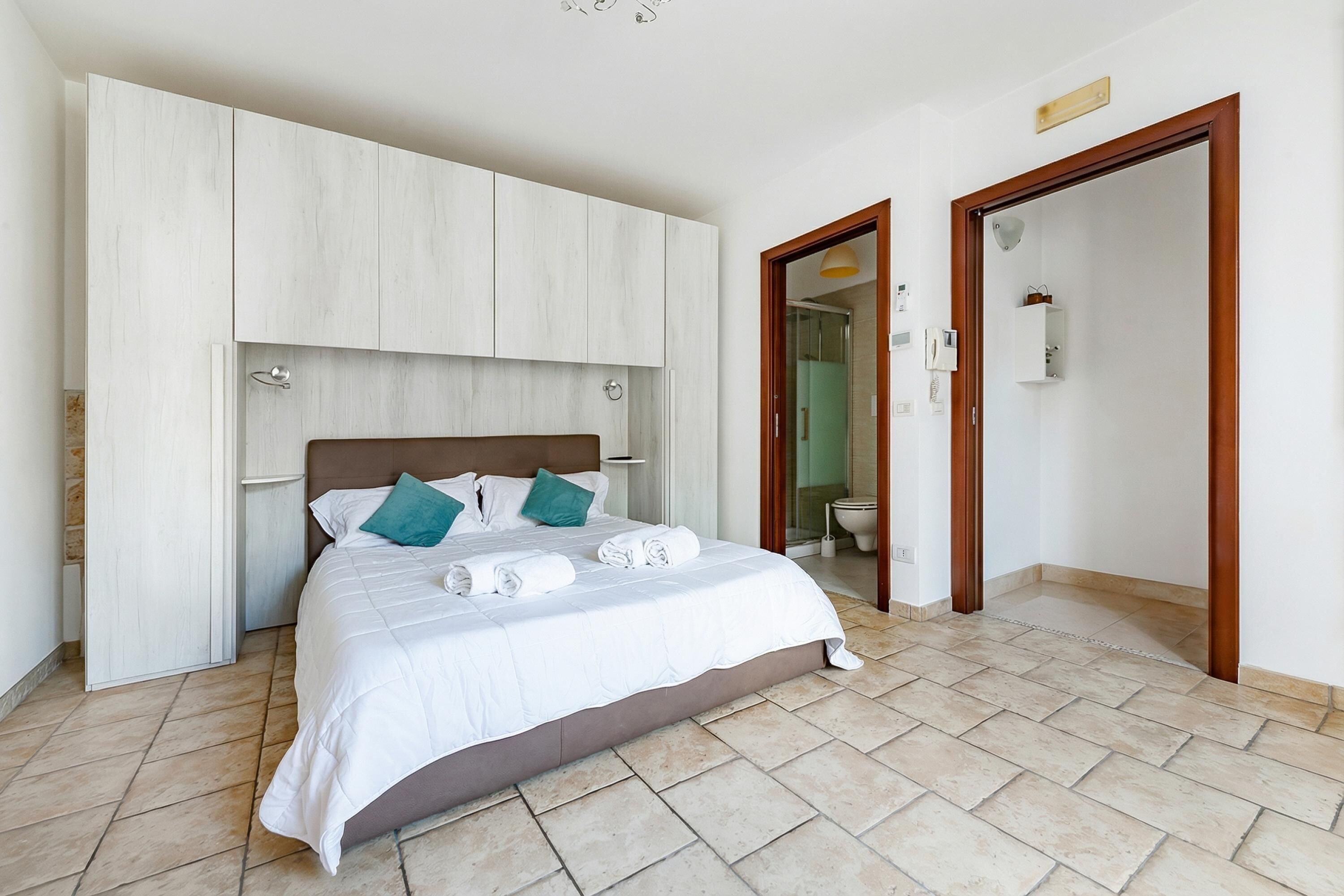 Een slaapkamer met een bed, witte wanden, een houten hoofdbord, een badkamers deur en een betegelde vloer.
