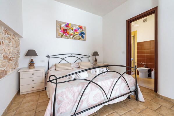 A bedroom with a bed, bedside tables, a painting, and a bathroom visible through an open door. - Helios Room (Conversano)