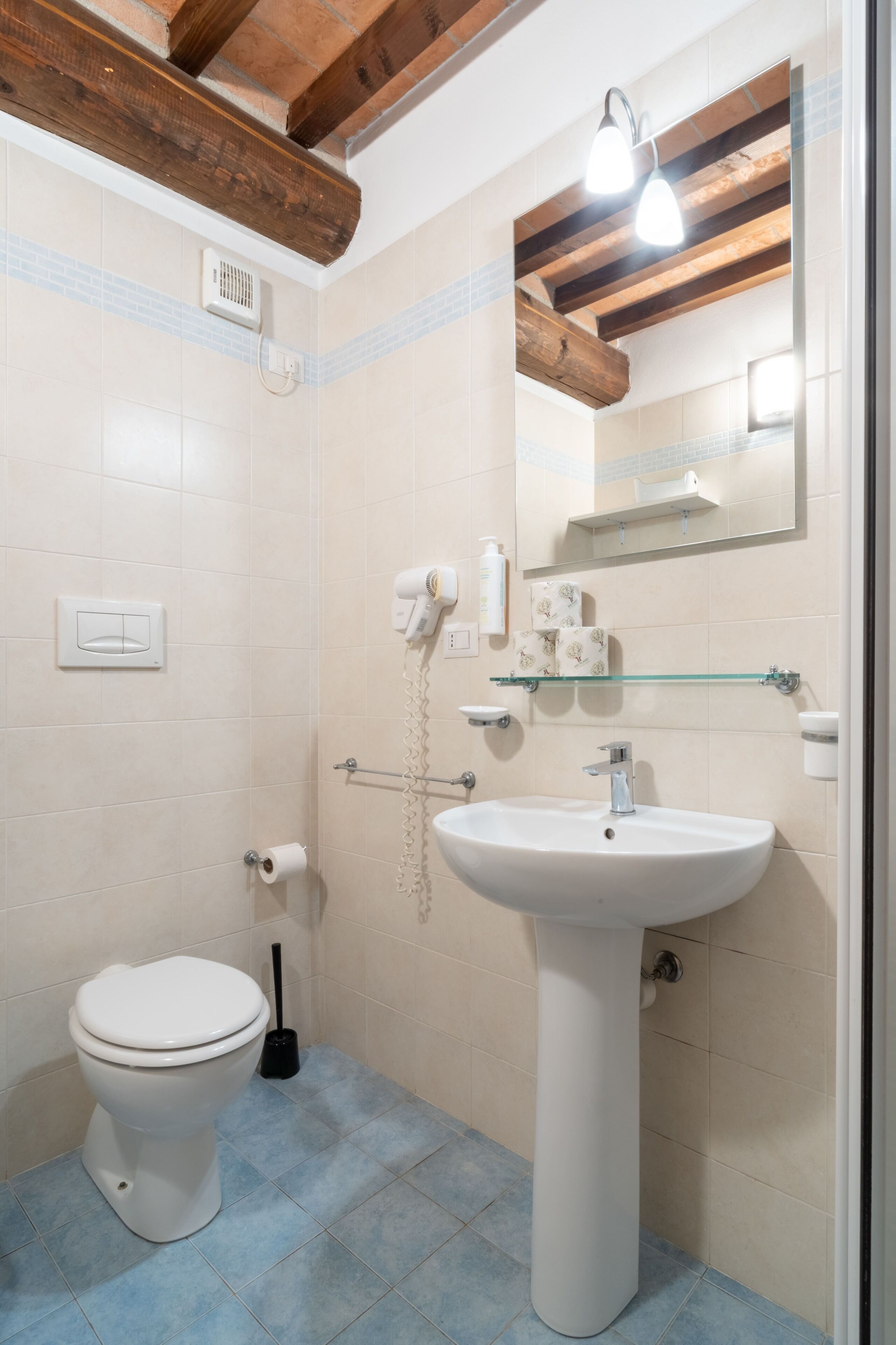 A bathroom with a white toilet, a pedestal sink, a glass shelf, a mirror, and a ceiling fan.