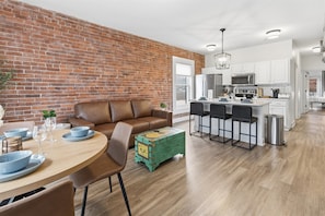 A modern kitchen with white cabinets, a central island, and a dining area with a brown leather sofa and a wooden table.
