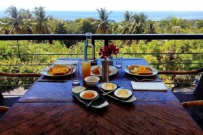 A dining table set with plates, bowls, and a glass of juice, overlooking a tropical landscape with palm trees and the ocean. - Panorama de Argao Boutique Resort (Argao)