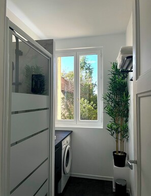 A compact laundry room with a washing machine, a drying rack, and a window providing a view of greenery outside. - Elegance and comfort at the gateway to Paris (Aulnay-sous-Bois)