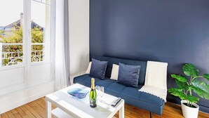 A modern living room with a dark blue sofa, white cushions, a white coffee table with a bottle and glasses, and a large window with a view of trees and buildings. - Elegance and comfort at the gateway to Paris (Aulnay-sous-Bois)