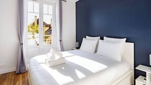 A modern bedroom with a large bed, white bedding, a dark blue accent wall, and a view through a window with curtains. - Elegance and comfort at the gateway to Paris (Aulnay-sous-Bois)