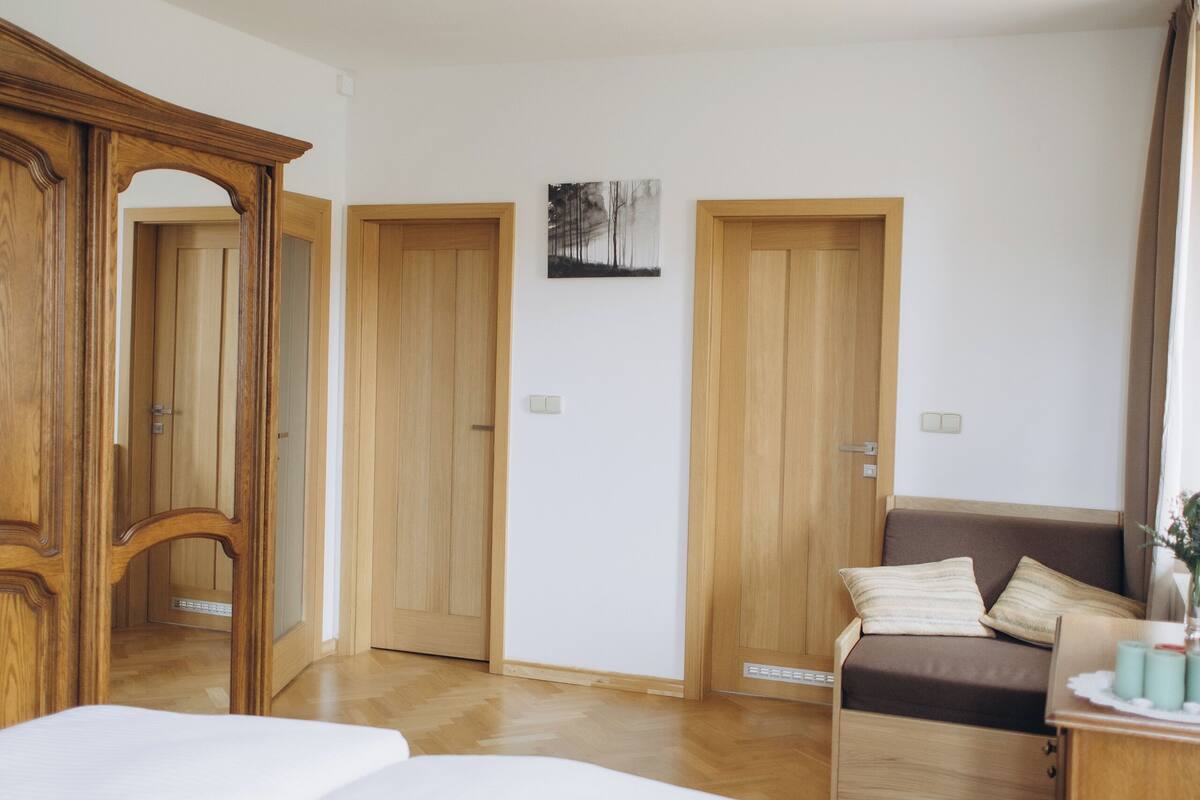A bedroom with a large bed, a wooden wardrobe, a bedside table, and a door leading to another room.