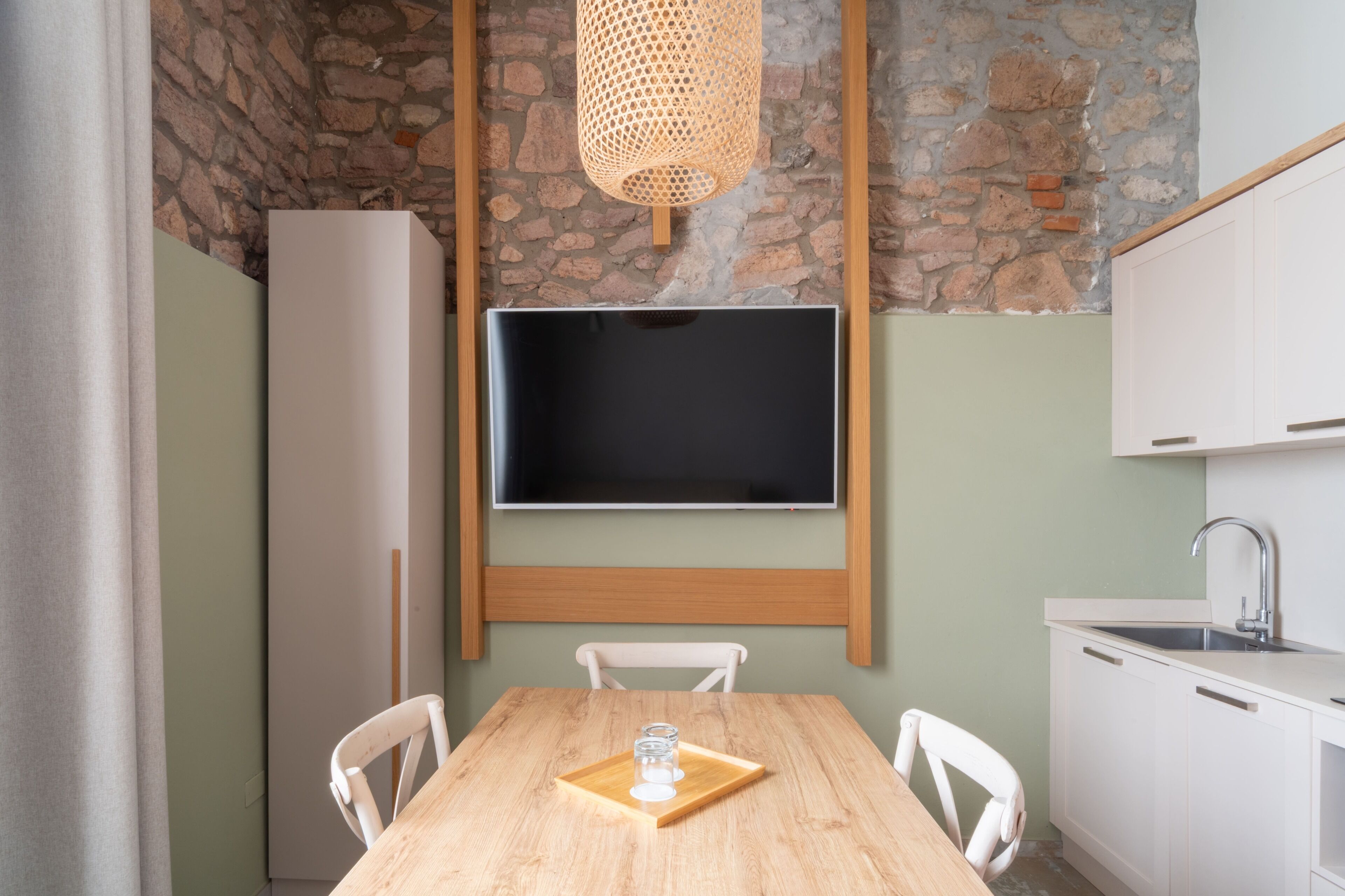 A kitchen with a stone wall, a wooden table, white chairs, a white refrigerator, and a mounted flat-screen TV.