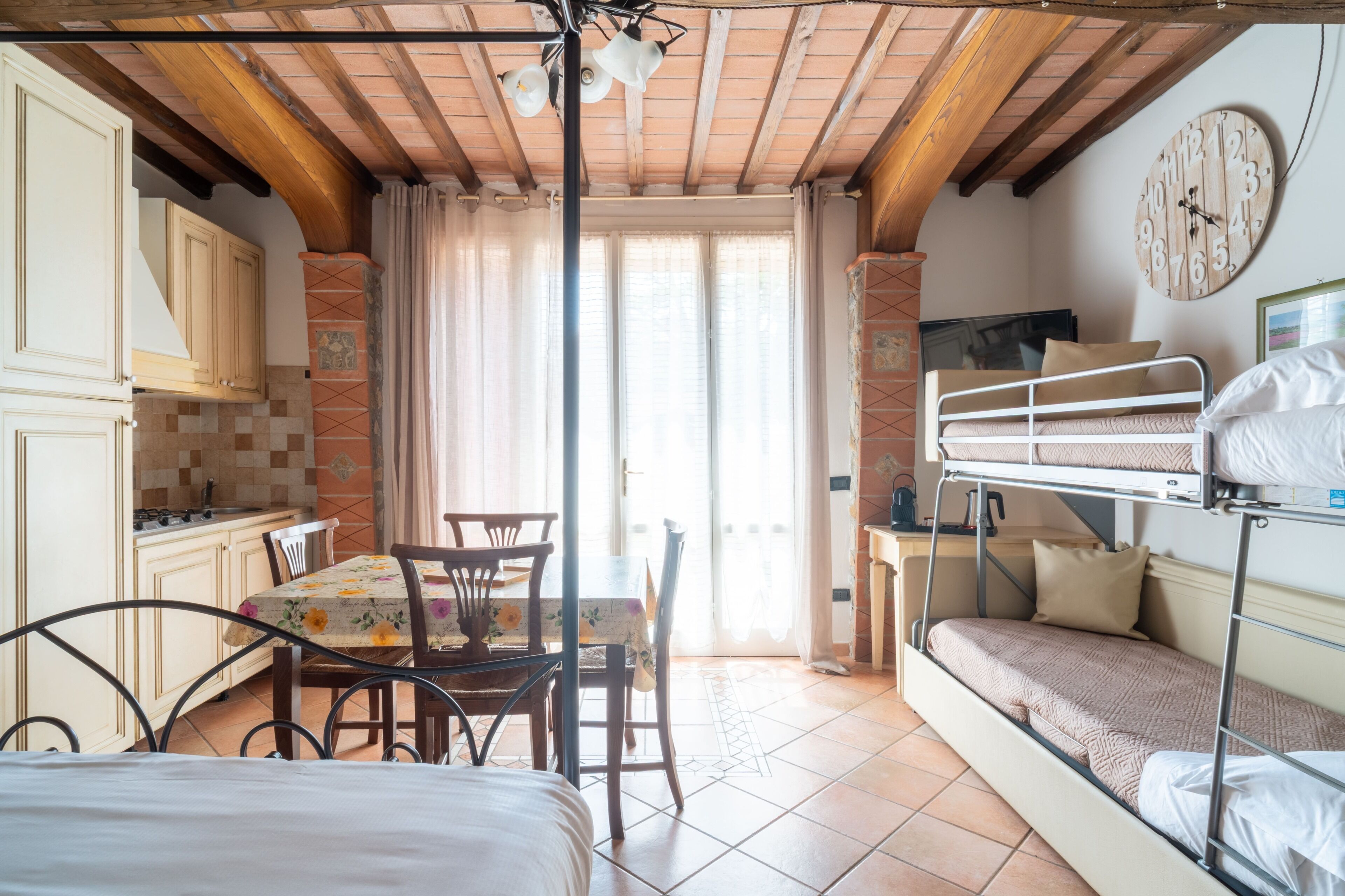 Una habitación con literas, una mesa de comedor con sillas, una zona de cocina y un reloj de pared grande.
