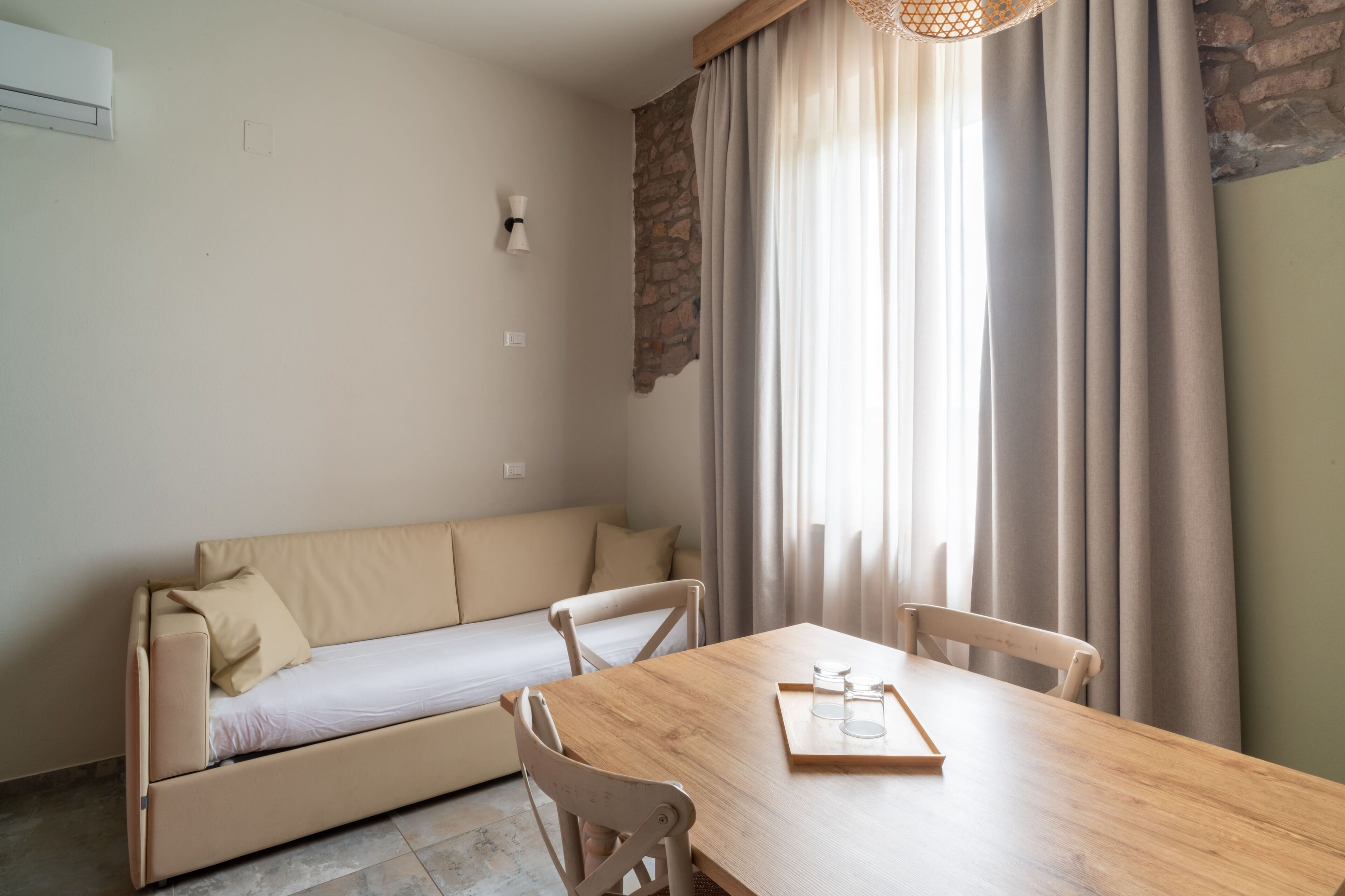 Una habitación con sofá, mesa de madera para comer, sillas, una bandeja con vasos y una ventana con cortinas.