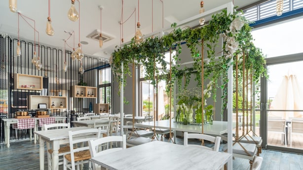 A restaurant with white wooden tables and chairs, hanging light bulbs, and green vines. - Hotiday Relais Costa degli Etruschi (Rosignano Marittimo)