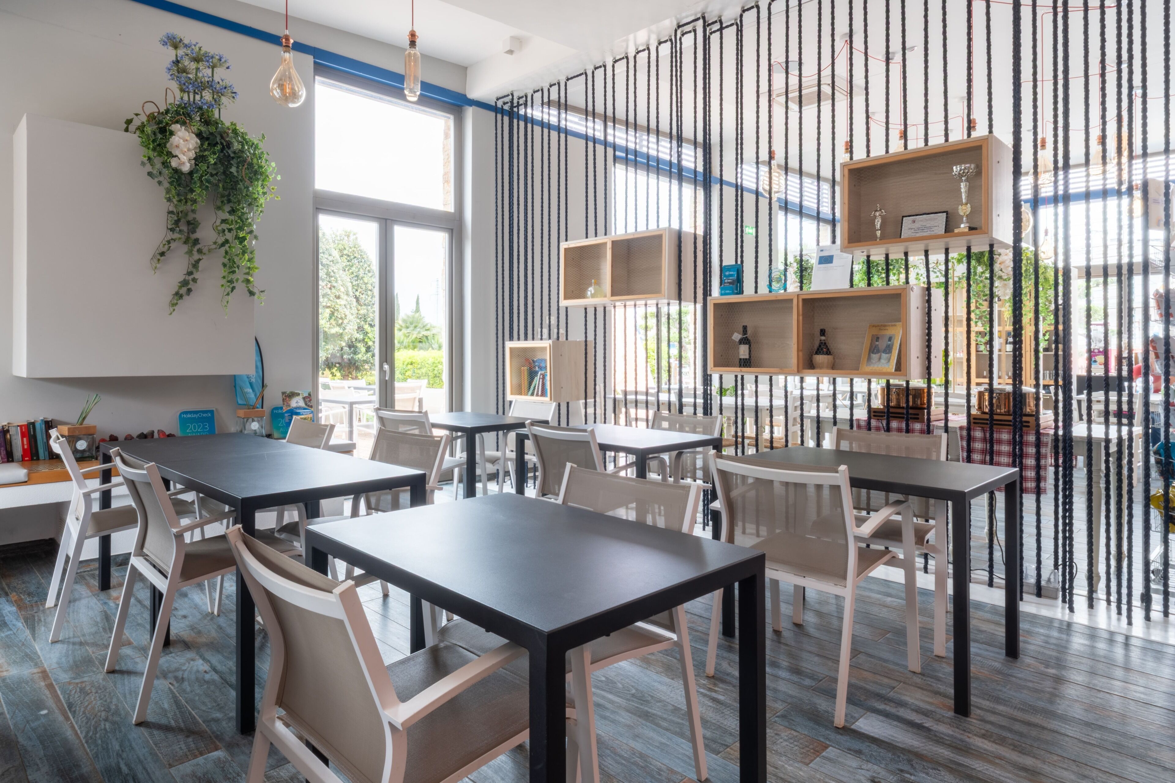A modern dining area with tables and chairs, a hanging plant, and shelves with items.