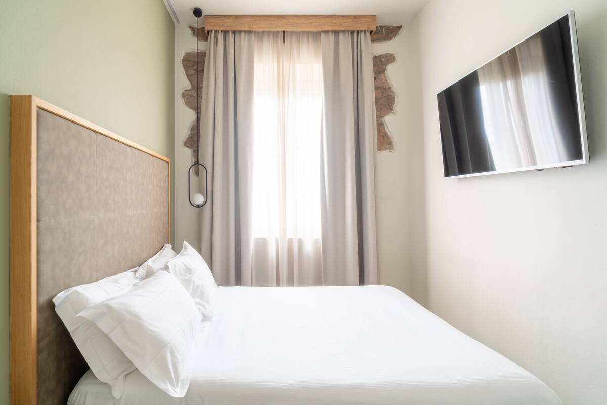 A hotel room with a bed, a TV mounted on the wall, and a window with curtains.