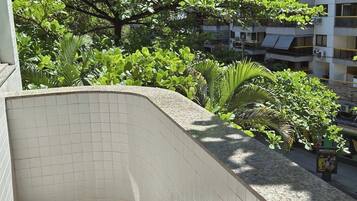 Uma área externa com piso de azulejos e um muro curvo, rodeada por vegetação exuberante e um edifício alto ao fundo.