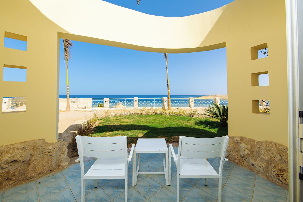 Een terras met twee witte stoelen en een tafel, met uitzicht op een strand met een helderblauwe lucht en oceaan.