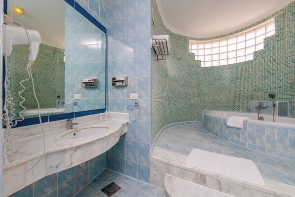 A bathroom with a marble sink, a large mirror, a built-in bathtub, and a window providing natural light.