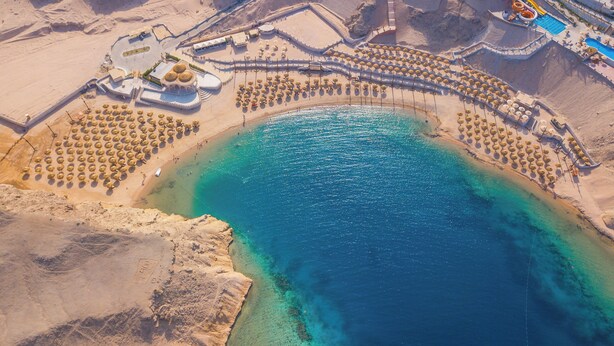 Vista aérea de un complejo turístico frente al mar, con agua azul cristalina, playas de arena y una zona de piscina.