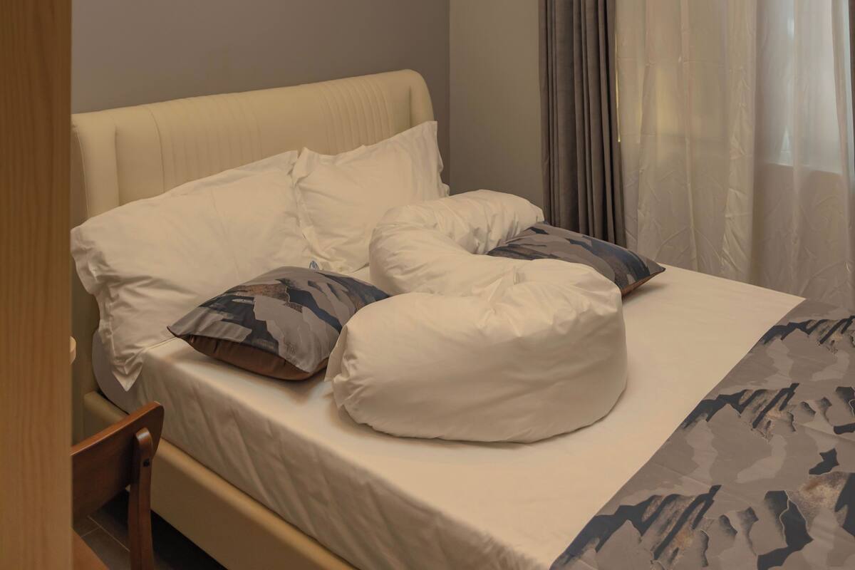 A neatly made bed with white linens and multiple pillows, a wooden nightstand, and a ceiling light fixture.