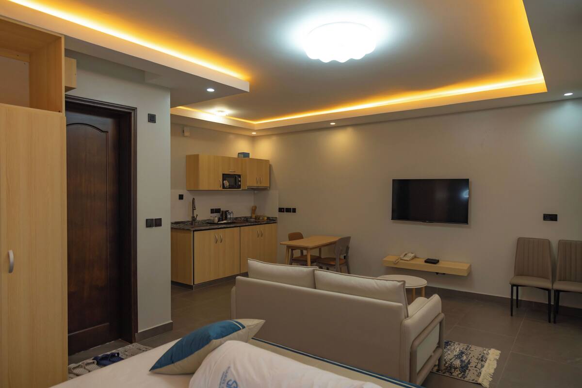 A modern hotel room with a bed, a sofa, a TV, a small kitchen area, and a dining table.