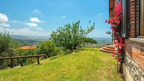 Ein grasbewachsener Hügel mit einer hölzernen Bank, ein Backsteinhaus mit roten Blumen und Blick auf ferne Hügel und Gebäude.