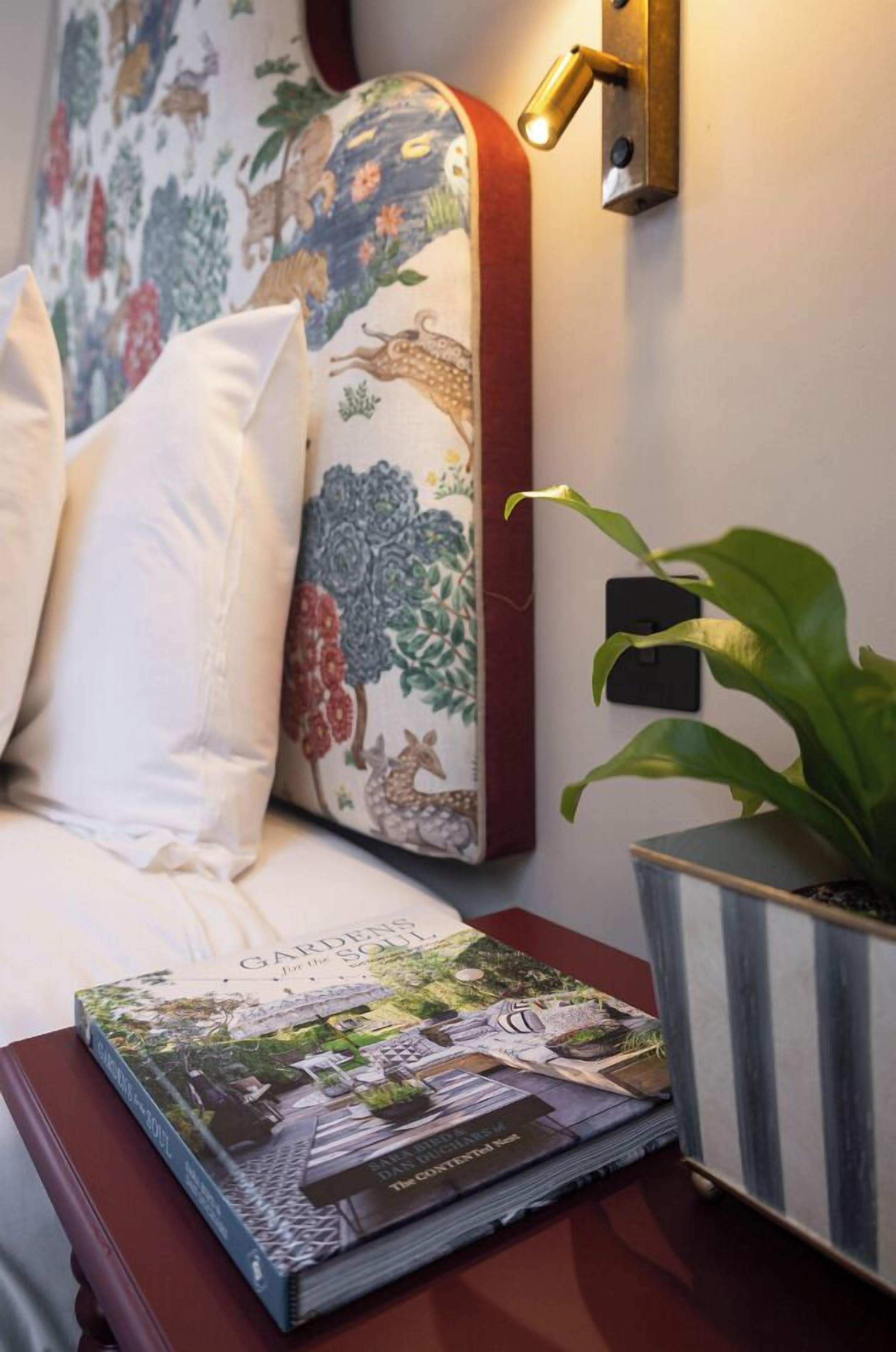 A neatly made bed with white linens, a decorative pillow with floral patterns, a book titled 'Garden's Soul', and a potted plant on a bedside table.