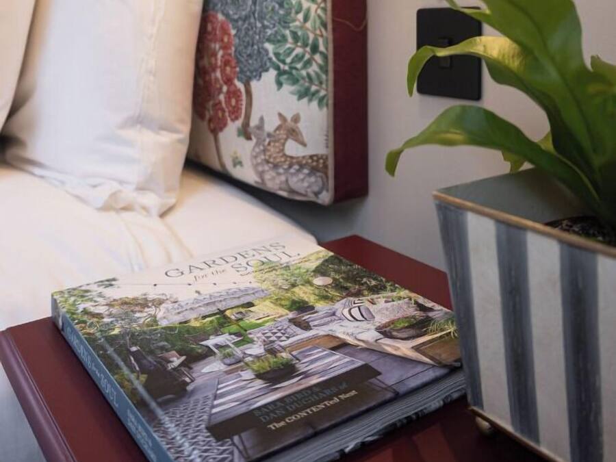 A neatly made bed with white linens, a decorative pillow with floral patterns, a book titled 'Garden's Soul', and a potted plant on a bedside table.