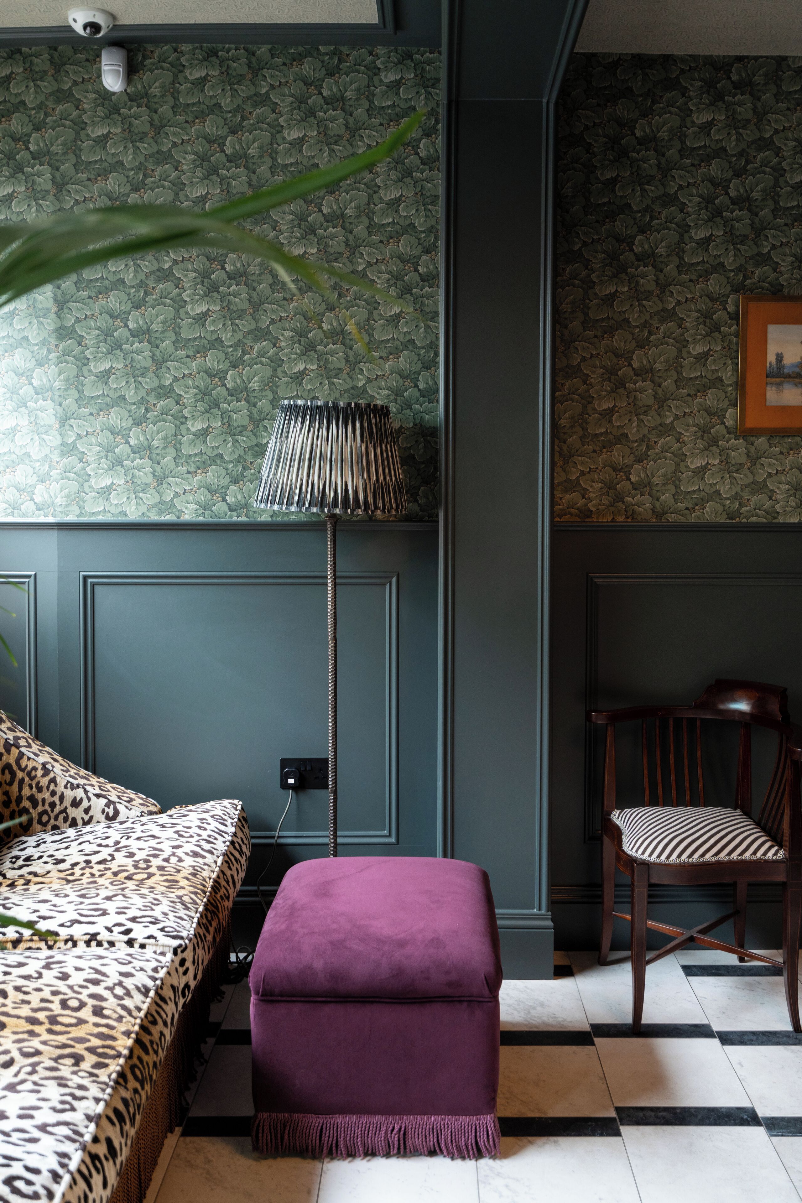 Una habitación con papel tapiz estampado, una cama con estampado de leopardo, un reposapiés morado, una silla con cojín a rayas y una lámpara de pie.