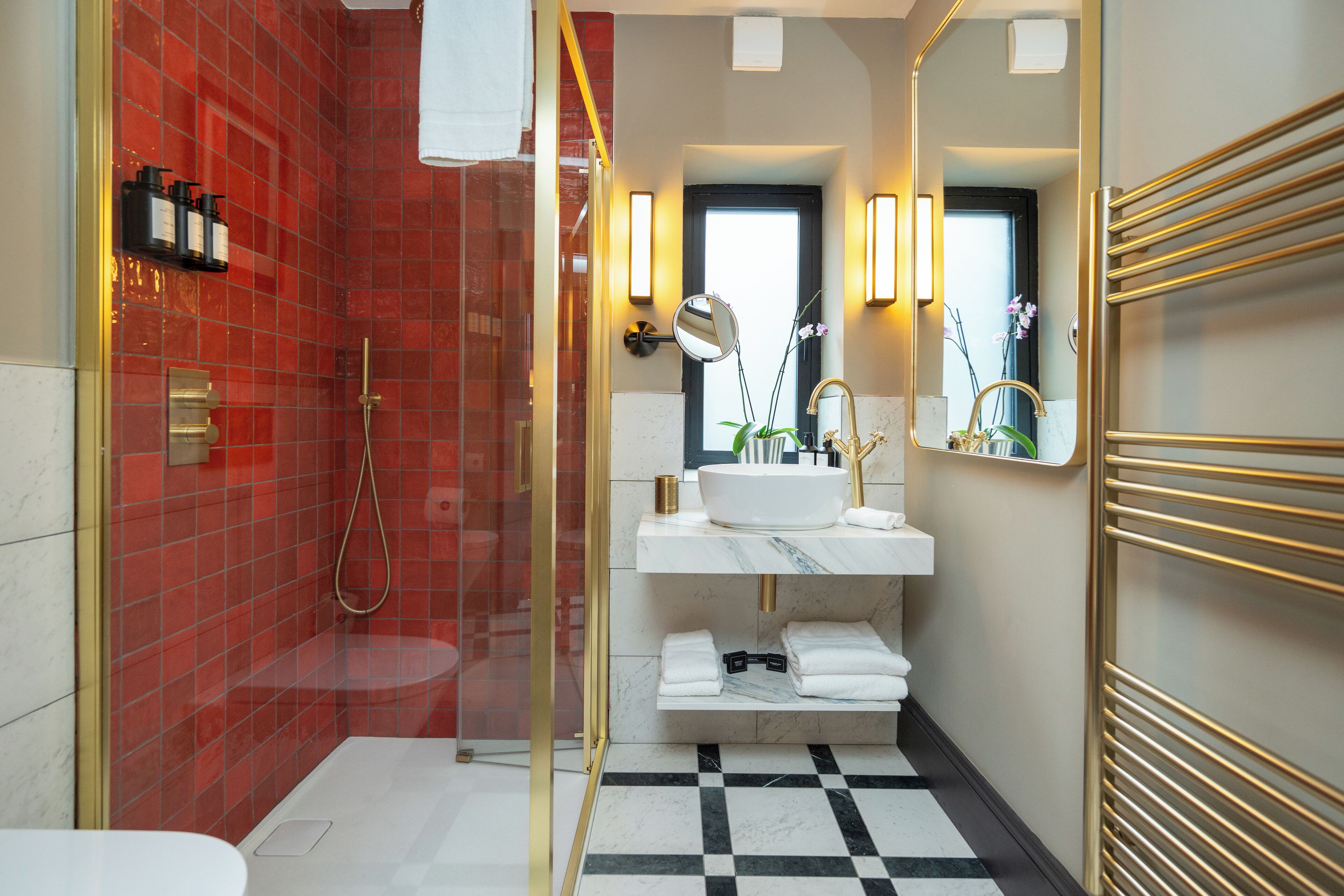 A modern bathroom with a red-tiled shower, a white sink with gold fixtures, and a black and white checkered floor.