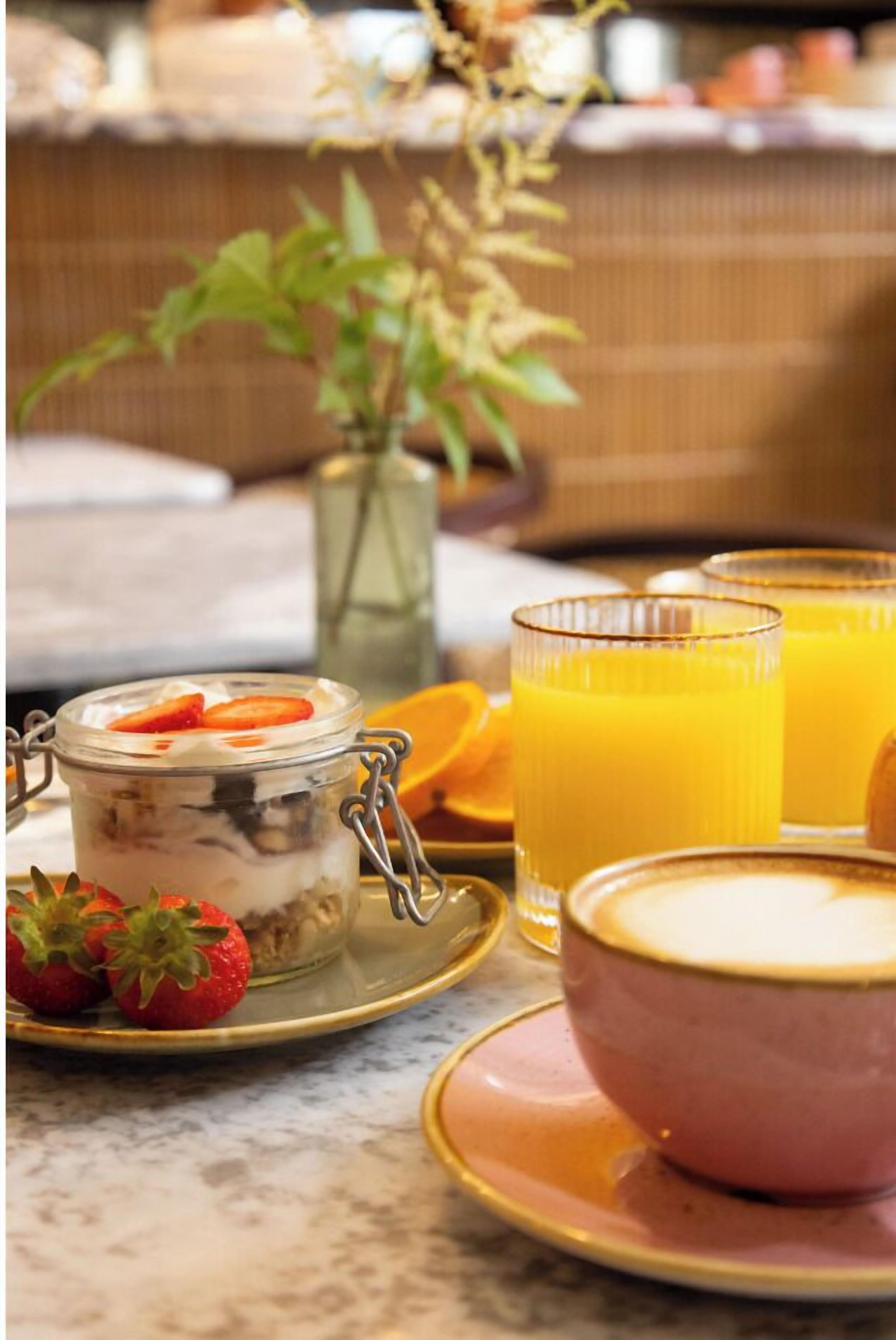 Un desayuno con un tarro de postre en capas, fresas frescas, un vaso de zumo de naranja y un bol de sopa sobre una mesa de mármol.