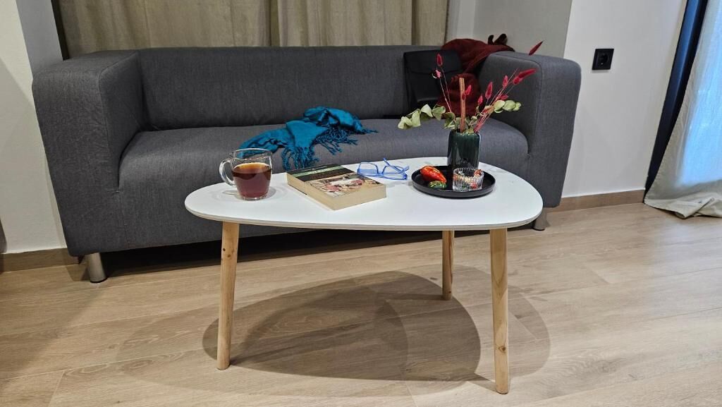 A modern living room with a grey sofa, a white coffee table with wooden legs, a book, a glass of tea, and a decorative vase with flowers.