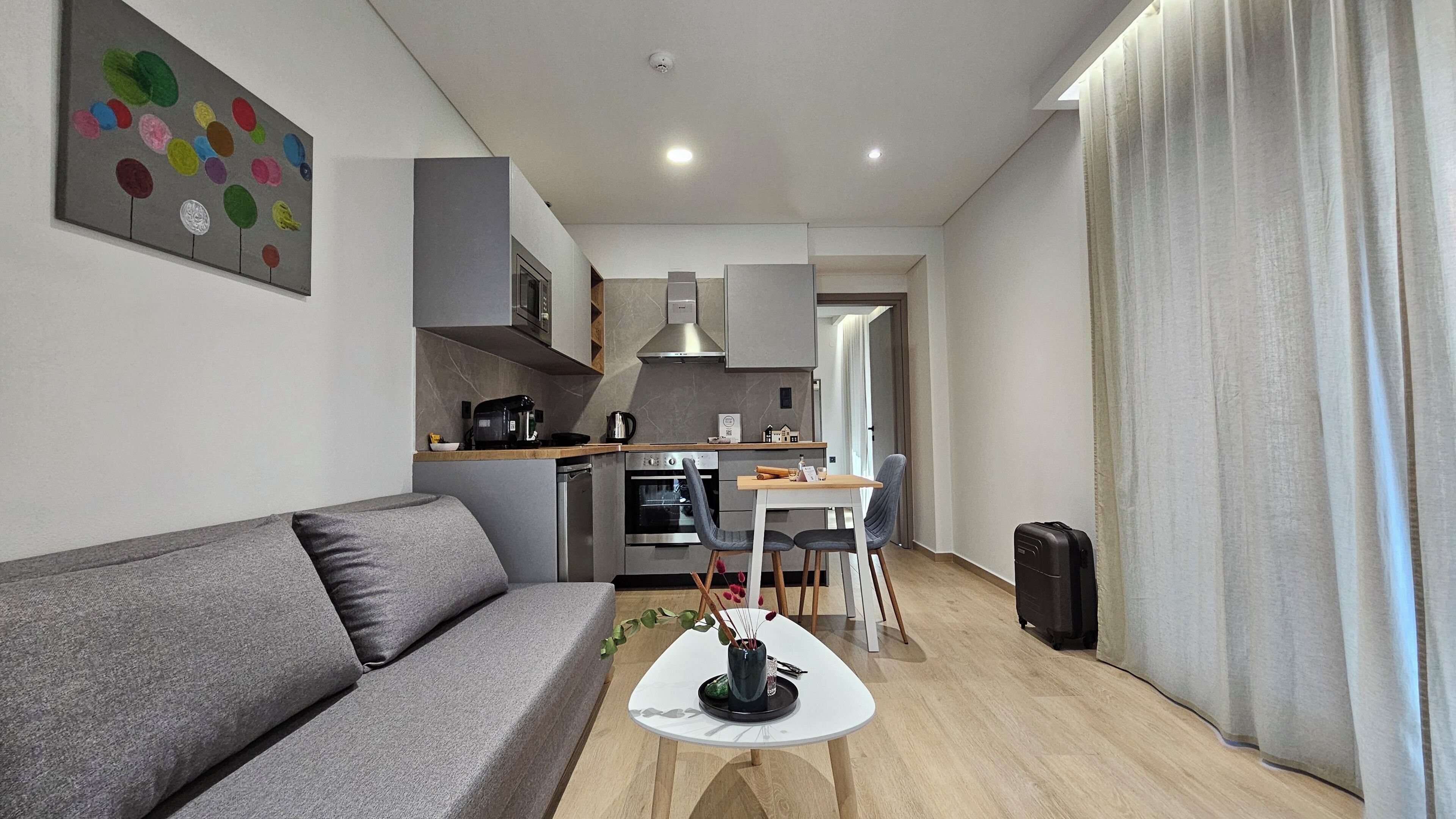 Ein modernes Wohnzimmer mit einem grauen Sofa, einem kleinen weißen Tisch mit einer Vase und einer Küchenzeile mit Mikrowelle, Backofen und Kaffeemaschine.