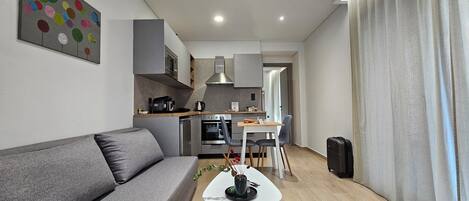Ein modernes Wohnzimmer mit einem grauen Sofa, einem kleinen weißen Tisch mit einer Vase und einer Küchenzeile mit Mikrowelle, Backofen und Kaffeemaschine.
