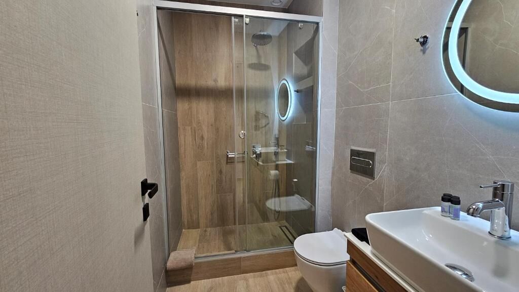 A modern bathroom with a glass-enclosed shower, a toilet, a sink with a mirror, and a wall-mounted light fixture.