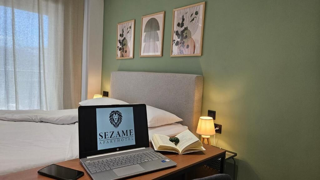 Ein Hotelzimmer mit einem Bett, einem Laptop, der das Logo 'SEZAME APARTHOTEL' zeigt, einer Nachttischlampe, einem Buch und einem Smartphone auf dem Nachttisch.