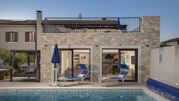 Ett poolområde med solstolar och parasoller, en byggnad i sten med en balkong och en klar himmel.