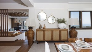 A modern dining area with a wooden dining table set for four, a wicker sideboard, and a view of the city through large windows. - Luxurious Villa Rizes - with Private Pool (Maroulas)