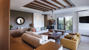 A modern living room with a sofa, coffee table, and chairs, wooden beams on the ceiling, and a large window with a view of trees. - Luxurious Villa Rizes - with Private Pool (Maroulas)