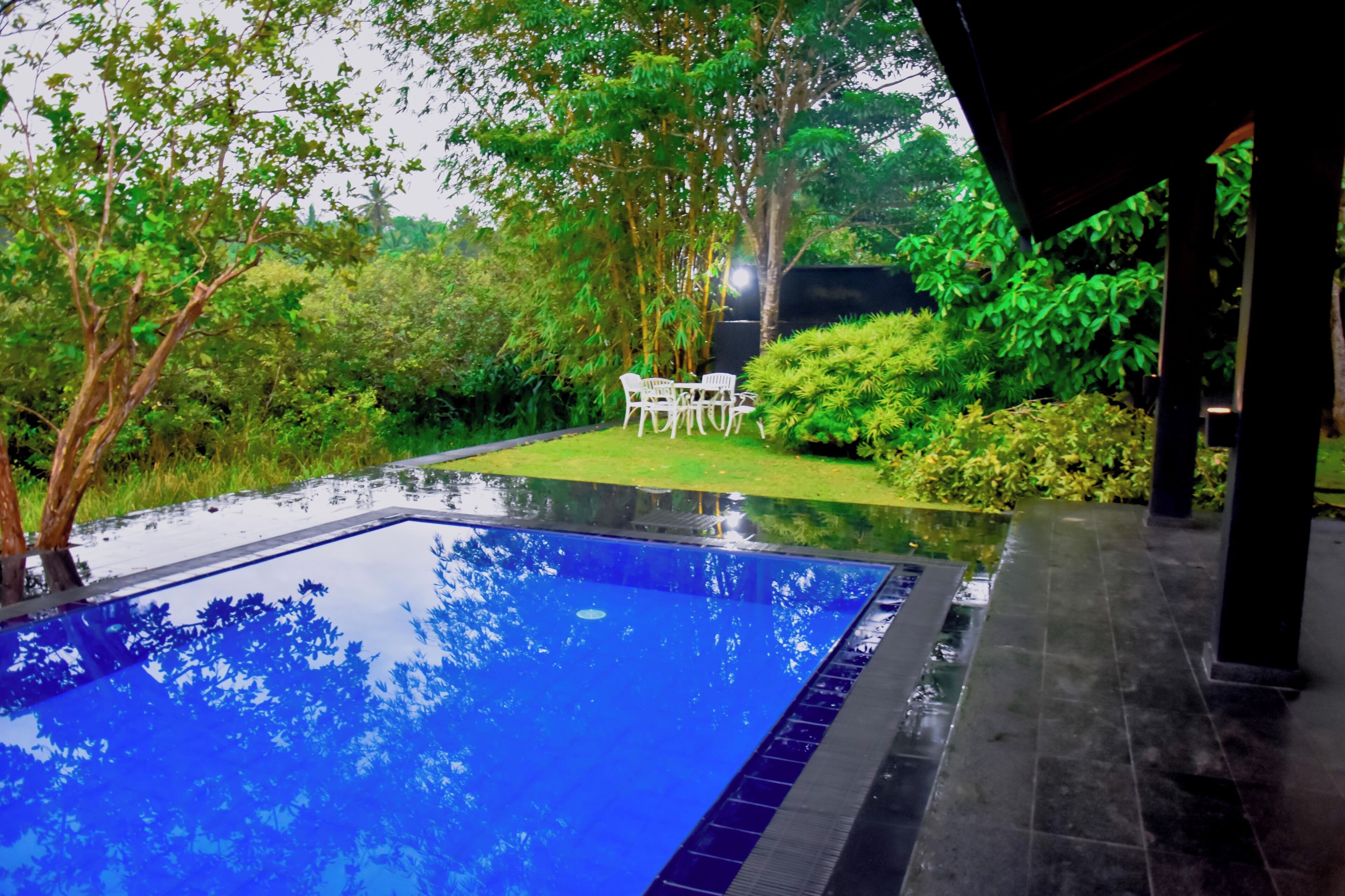 A swimming pool with a tiled deck, surrounded by lush greenery and a small table with chairs.