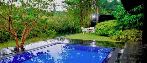 A swimming pool with a tiled edge, surrounded by lush greenery and a small white table with chairs.