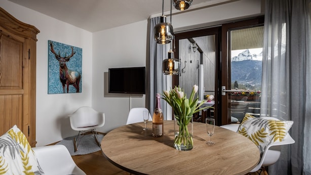 A dining area with a round wooden table, white chairs, a vase with flowers, and a painting of a deer. - Vacation apartment Bergkristall with Watzmann view (Berchtesgaden)